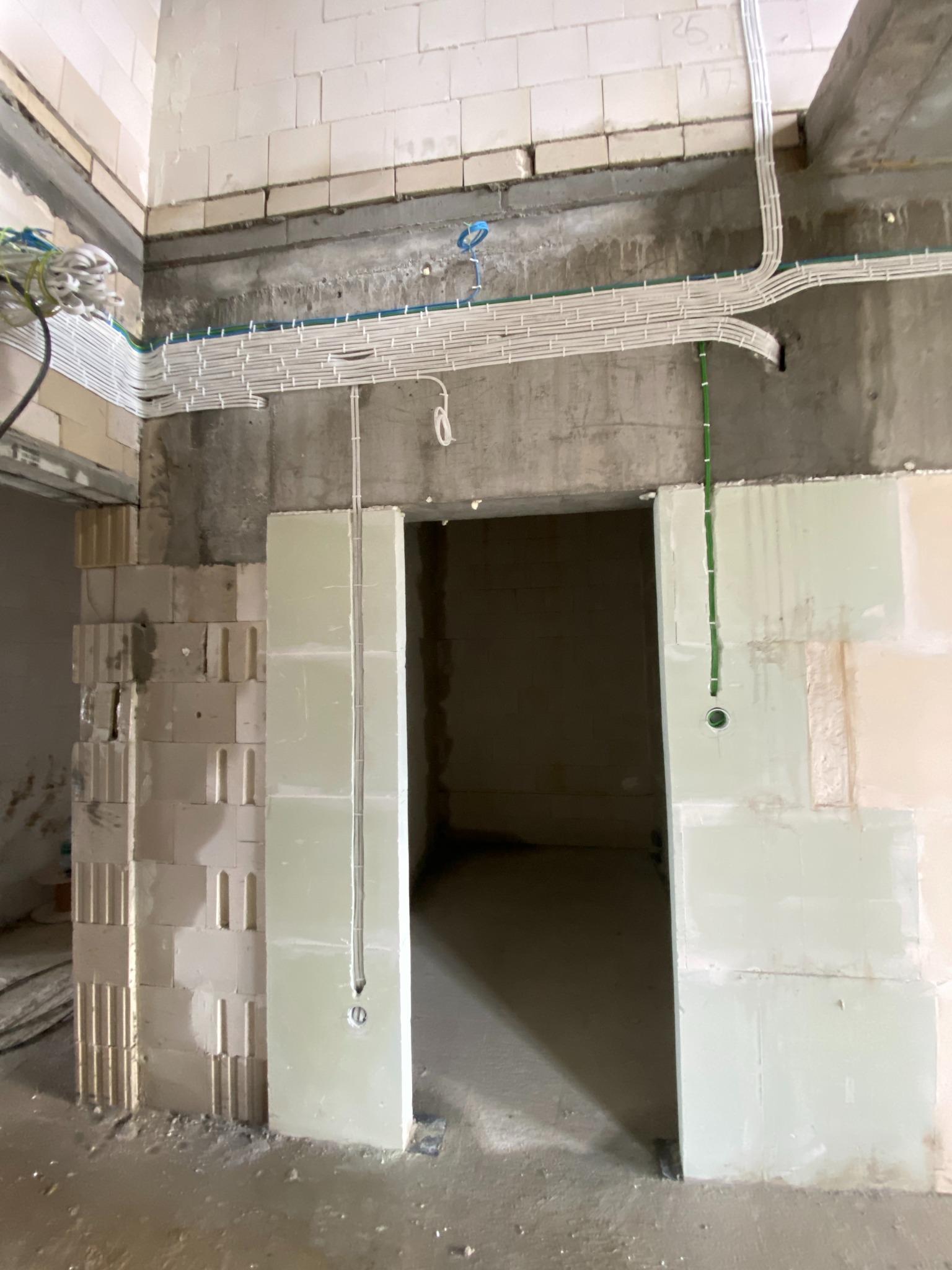 Surowe wnętrze w trakcie prac wykończeniowych z widocznymi przewodami elektrycznymi ułożonymi wzdłuż ściany i nad otworem drzwiowym, oraz pionowo w dół ściany, z przygotowanymi otworami pod gniazdka.