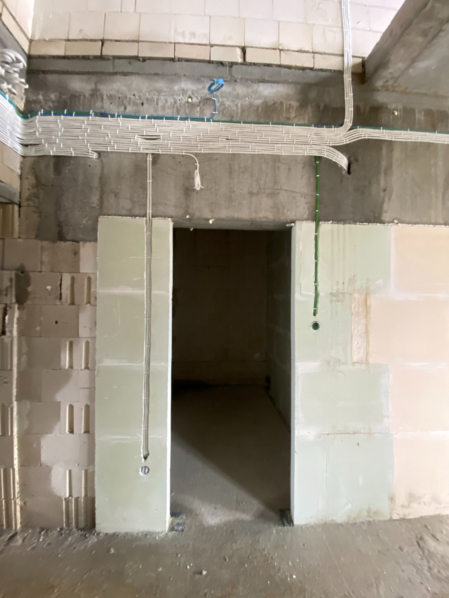 Surowy stan budynku w trakcie prac. Widoczne kable elektryczne ułożone pod sufitem i w ścianach, otwór drzwiowy w stanie deweloperskim, ściany z betonu komórkowego i pustaków.