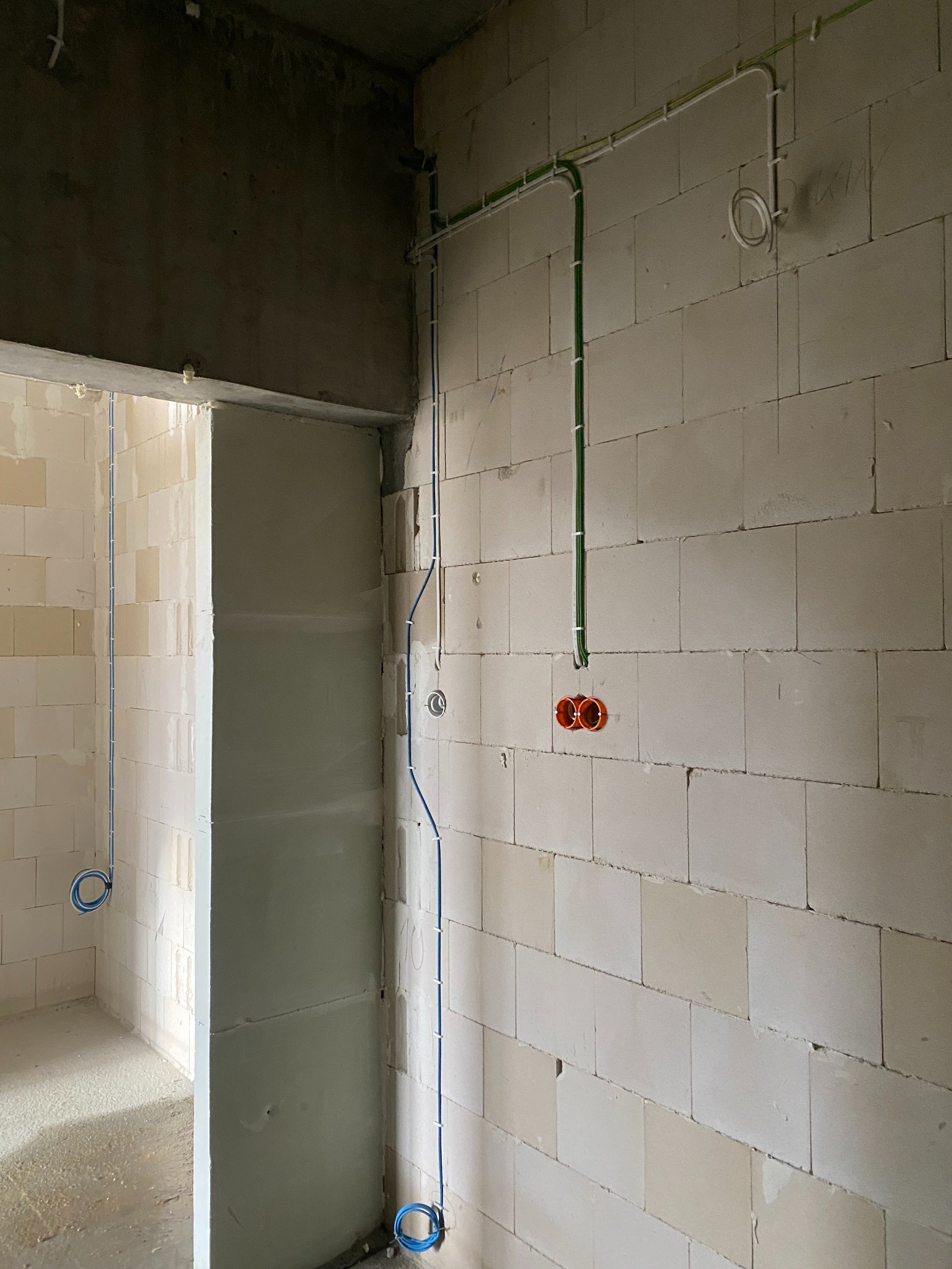 Wnętrze w stanie surowym: widoczne ściany z białych bloczków, pionowe kable elektryczne w kolorach niebieskim i zielonym, zamocowane do ściany za pomocą białych uchwytów, oraz pomarańczowe puszki...