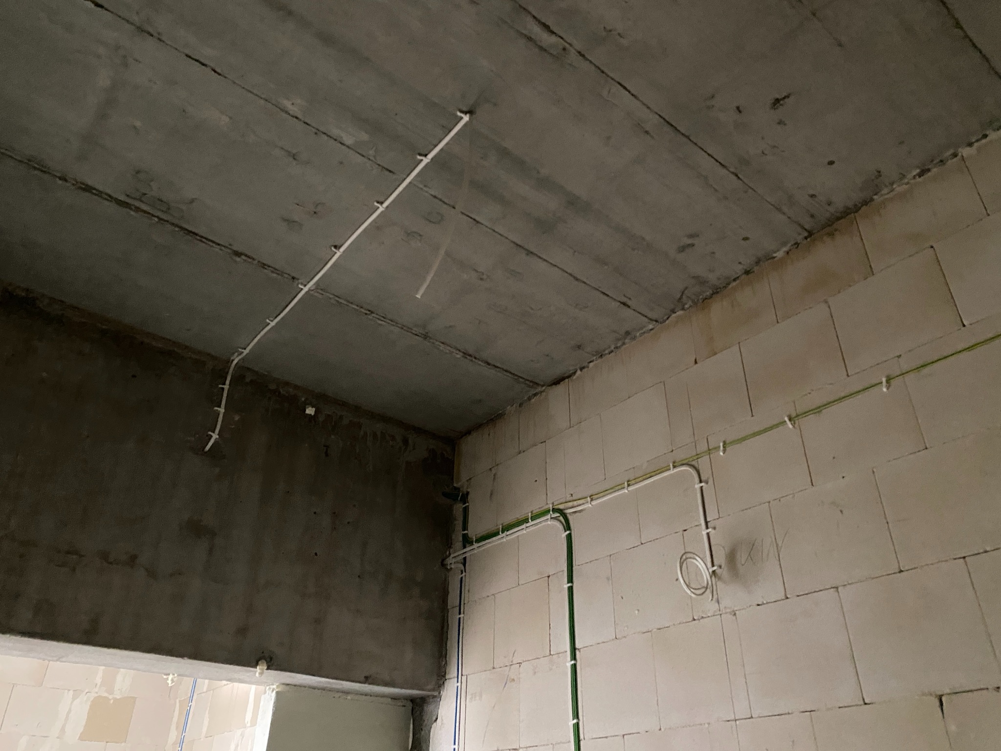 Surowy stan wnętrza: betonowy sufit z przymocowanymi białymi rurkami instalacji elektrycznej, ściany z jasnych bloczków z poprowadzonymi przewodami w peszlach, widoczne okablowanie.