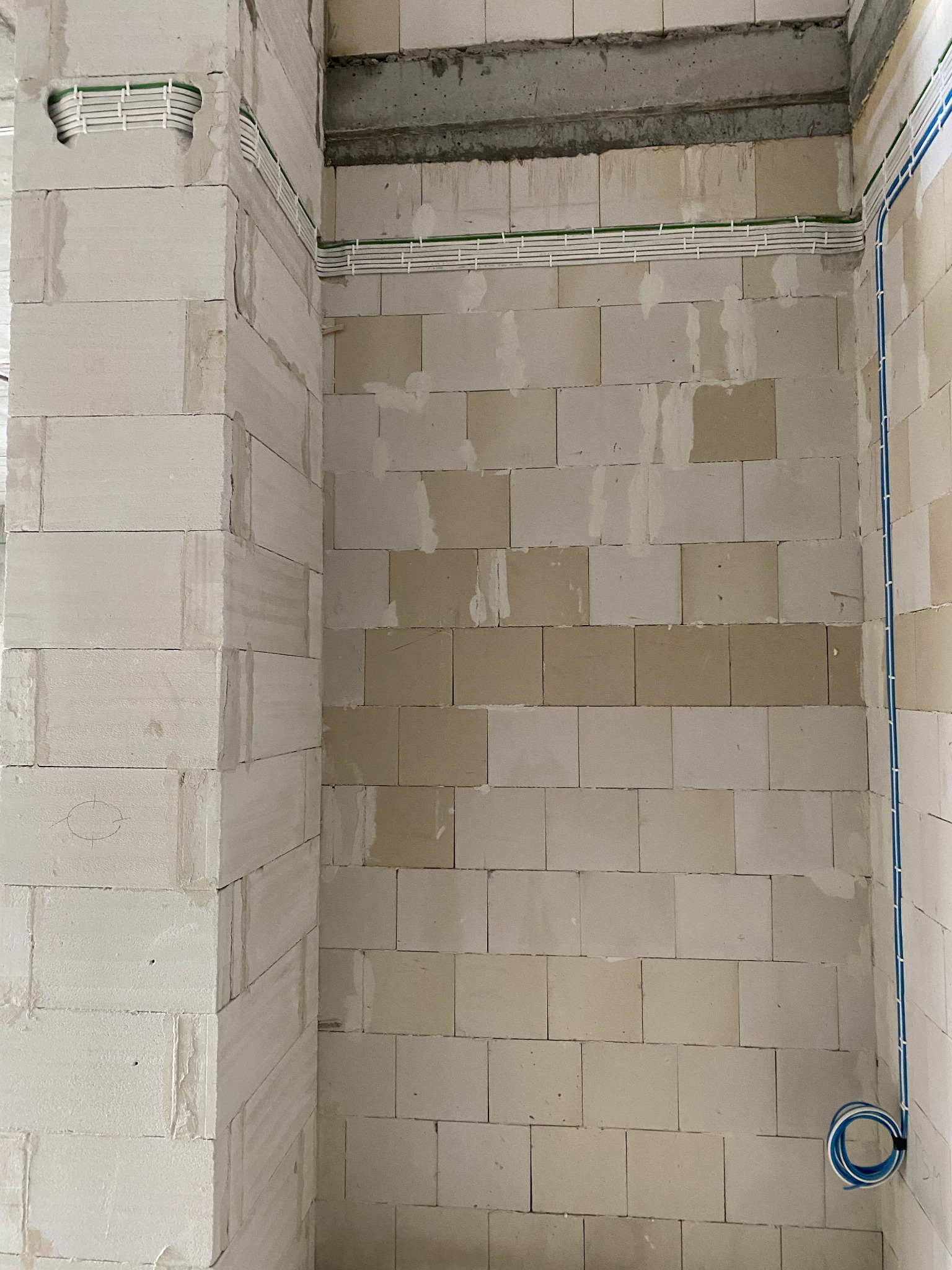 Ściana z betonu komórkowego w trakcie montażu instalacji elektrycznej. Widoczne białe korytka kablowe z zielonymi przewodami oraz niebieski przewód w peszlu, zwinięty w pętlę przy podłodze. Surowy...