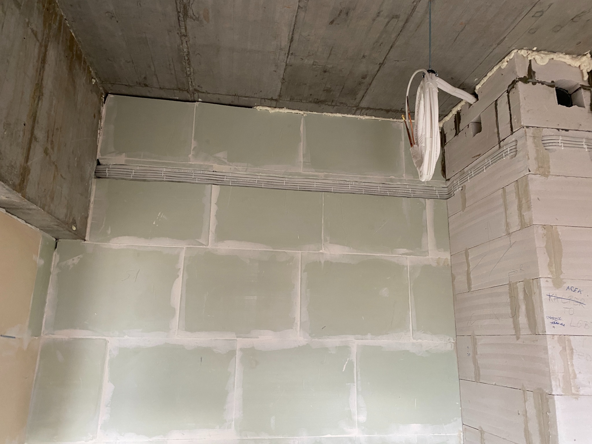 Wnętrze w trakcie remontu: ściany z płyt gipsowo-kartonowych, widoczne przewody w korytkach, surowy betonowy sufit i fragment ściany z betonu komórkowego z odręcznymi napisami.