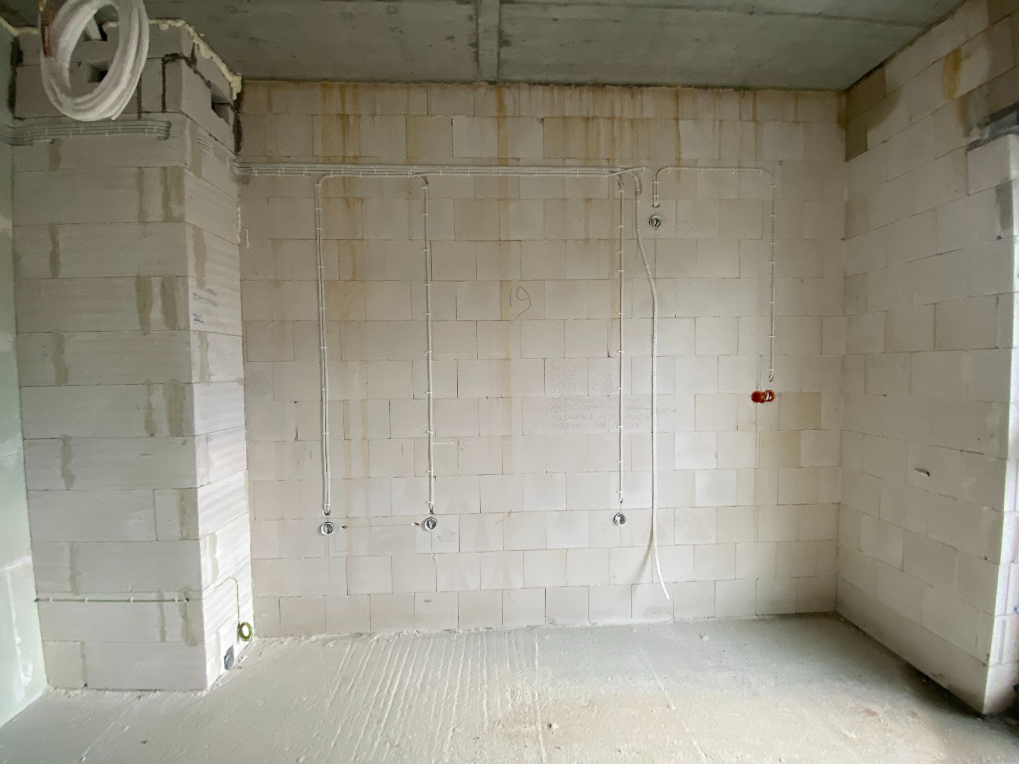 Surowy stan pomieszczenia z widocznymi przewodami elektrycznymi poprowadzonymi po ścianach z betonu komórkowego, z zamontowanymi puszkami pod gniazdka i wypustami kablowymi pod oświetlenie.