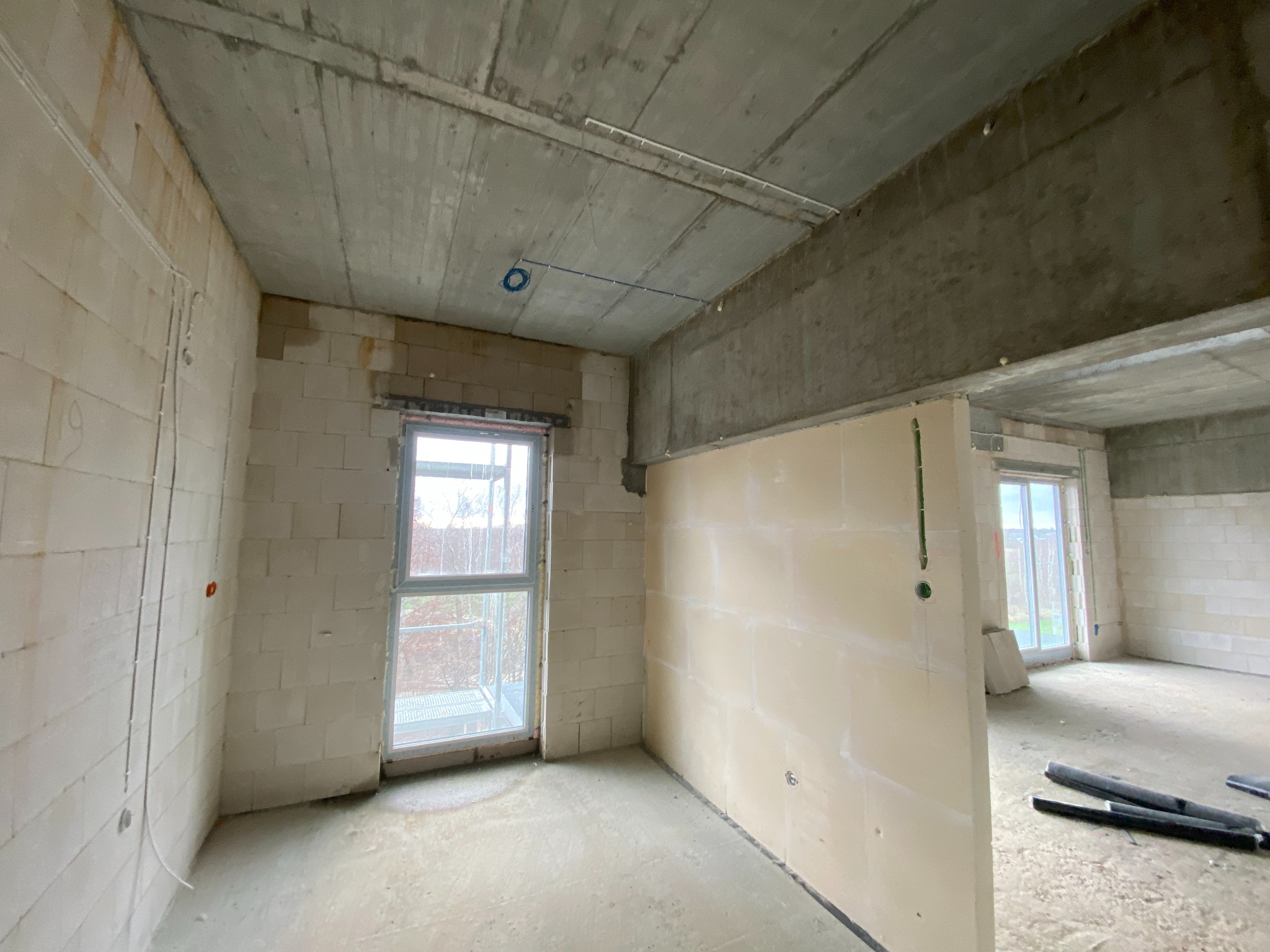 Surowy stan wnętrza budynku w trakcie prac wykończeniowych, widoczne instalacje elektryczne w ścianach i na suficie, ściany z bloczków i częściowo obłożone płytami gipsowymi, okno z widokiem...