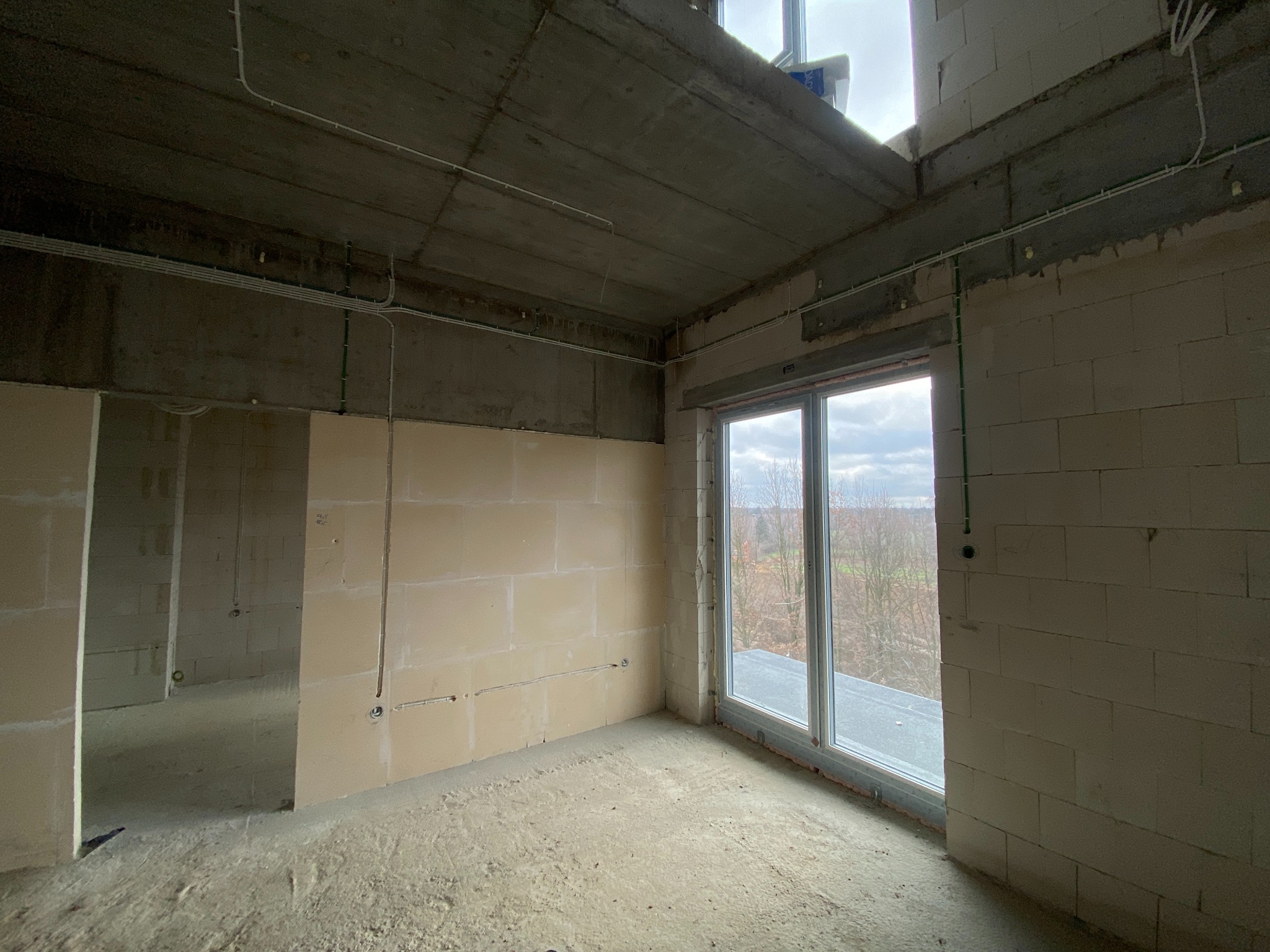 Surowy stan wnętrza z widocznymi przewodami elektrycznymi prowadzonymi po ścianach i suficie, okno z widokiem na zewnątrz, ściany z betonu komórkowego.