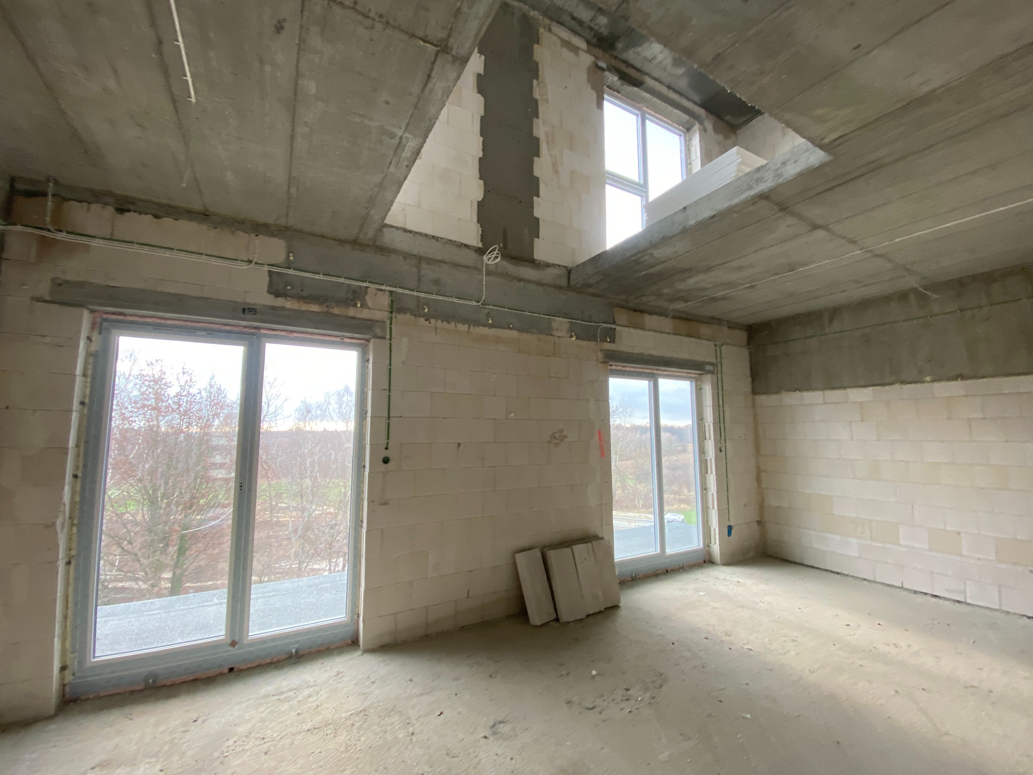 Surowy stan wnętrza budynku z widocznymi przewodami elektrycznymi pod sufitem, oknami i częściowo wykończonymi ścianami z betonu komórkowego.