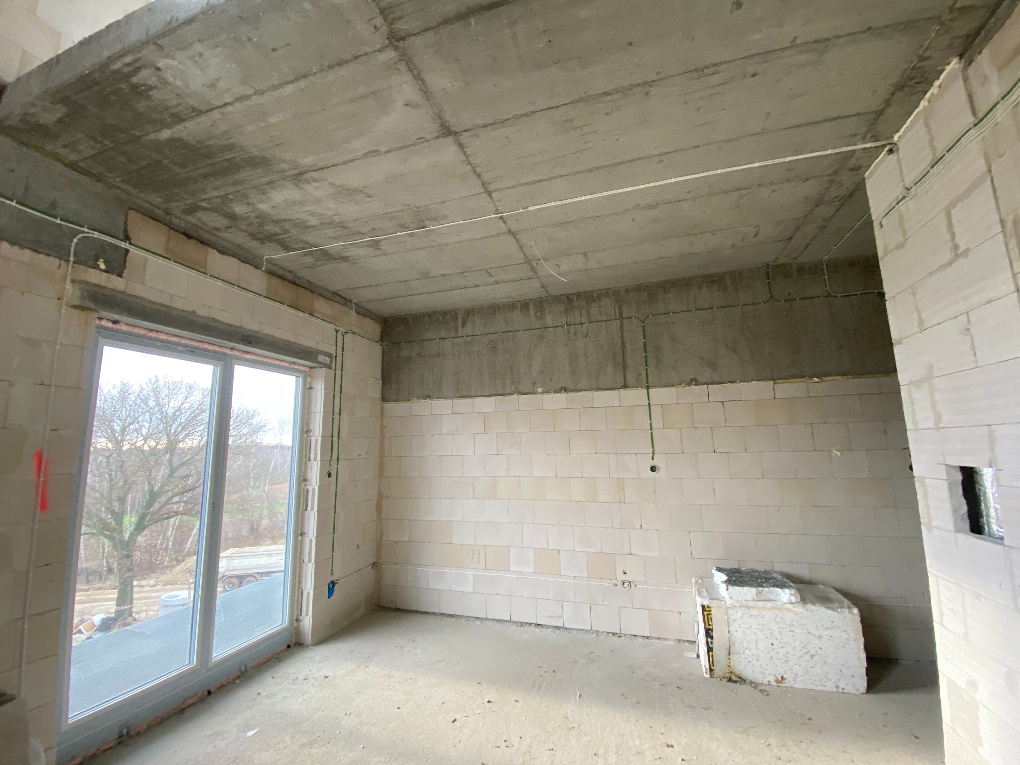 Surowy stan wnętrza w trakcie budowy, widoczne przewody elektryczne poprowadzone po ścianach z betonu komórkowego i pod sufitem, okno balkonowe z widokiem na zewnątrz.