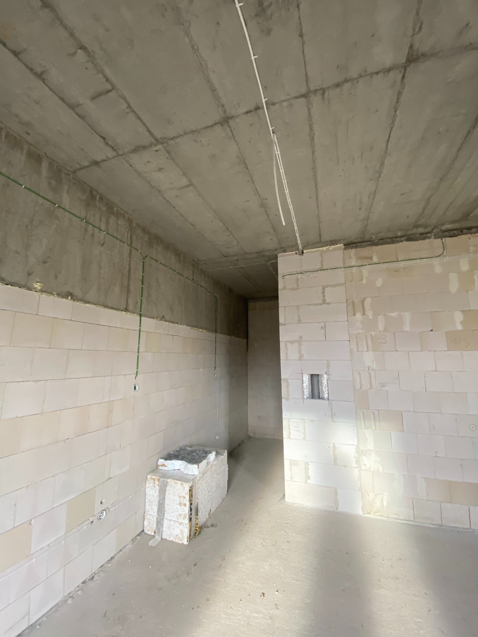 Surowy stan pomieszczenia z widocznymi przewodami elektrycznymi prowadzonymi po ścianach i suficie, ściany z białych bloczków, na podłodze resztki materiałów budowlanych.