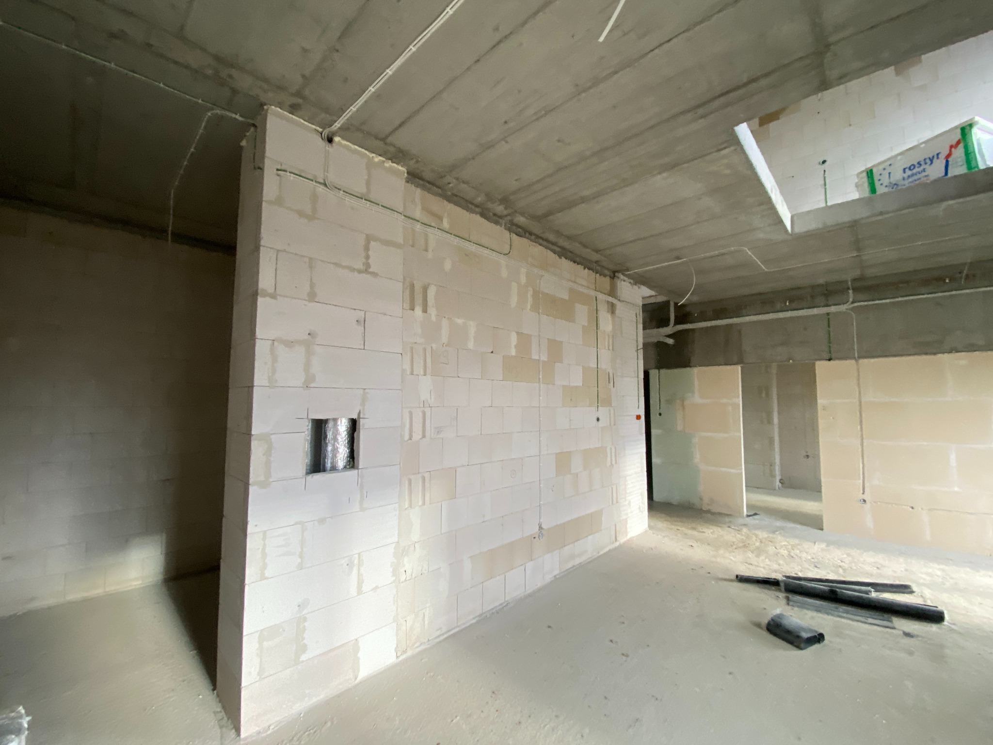 Surowy stan wnętrza w trakcie budowy z widocznymi przewodami elektrycznymi prowadzonymi po ścianach z betonu komórkowego, otworem wentylacyjnym w ścianie oraz rurami PCV leżącymi na podłodze.