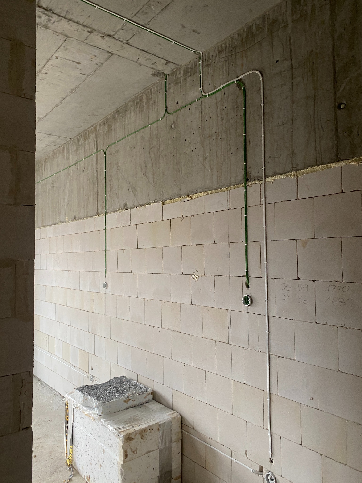 Wnętrze w stanie surowym: widoczne kable elektryczne prowadzone po ścianie z betonu komórkowego, z zaznaczonymi punktami pod gniazdka i włączniki. Na dole, przy ścianie, leżą bloczki budowlane.