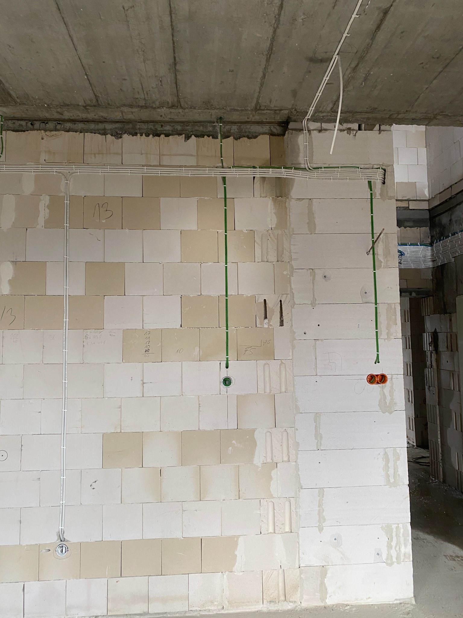 Surowy stan: widok na ścianę z betonu komórkowego z rozprowadzonymi przewodami elektrycznymi w peszlach i rurkach, widoczne puszki pod gniazdka i włączniki, sufit betonowy w tle, ściany z numerami...