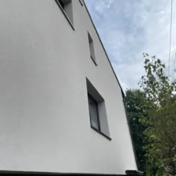 Świeżo otynkowana, jasnoszara elewacja domu jednorodzinnego z ciemnymi oknami, widoczna z dołu pod kątem, z fragmentem ogrodu w tle.