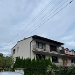 Dwupiętrowy dom z balkonami i jasną elewacją, widok z dołu, częściowo zasłonięty przez żywopłot i zaparkowany samochód dostawczy.