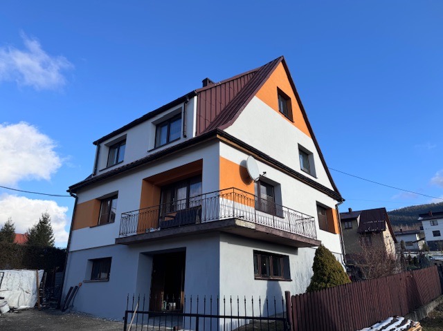 Dwupiętrowy dom w Ujsołach z balkonem i częściowo pomalowaną elewacją w kolorach białym i pomarańczowym, widoczny na tle błękitnego nieba.