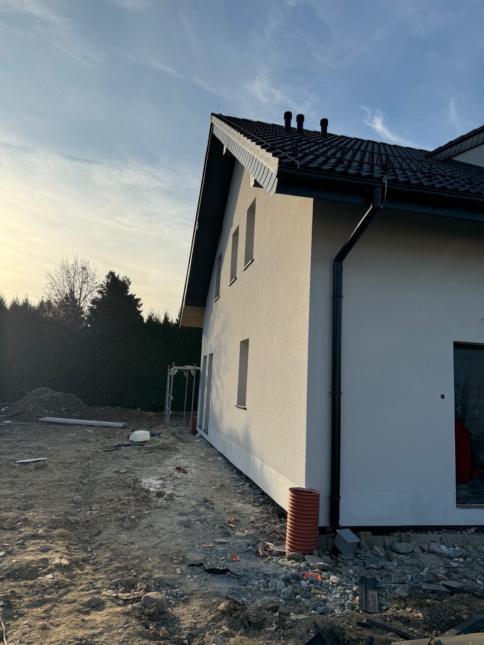 Świeżo otynkowana elewacja domu jednorodzinnego, widok z boku w porannym świetle, teren wokół jeszcze niezagospodarowany, widoczne rusztowanie i rura kanalizacyjna.