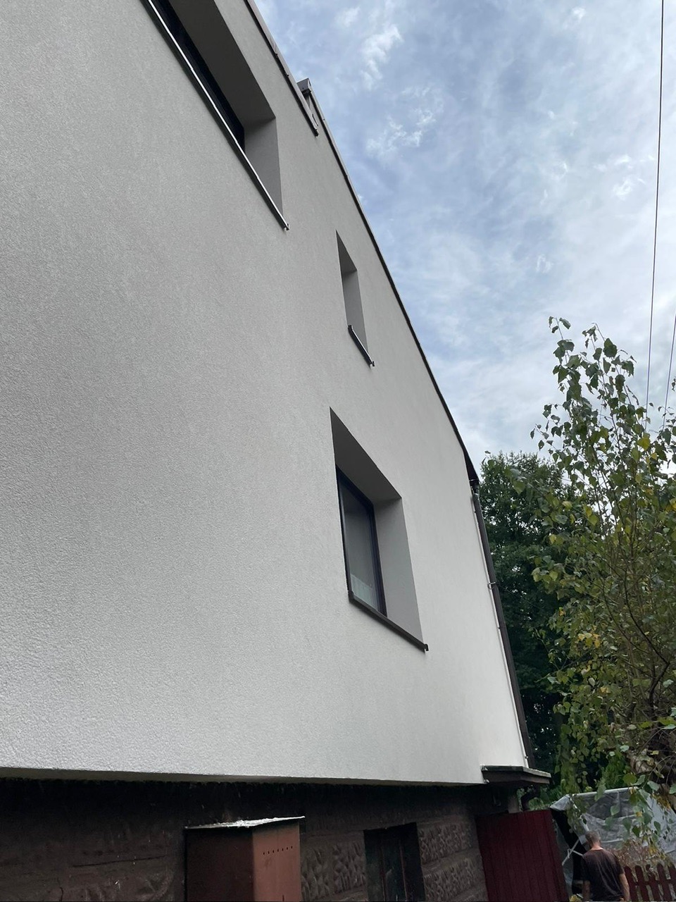 Świeżo otynkowana, jasnoszara elewacja domu jednorodzinnego z ciemnymi oknami, widoczna z dołu pod kątem, z fragmentem ogrodu w tle.