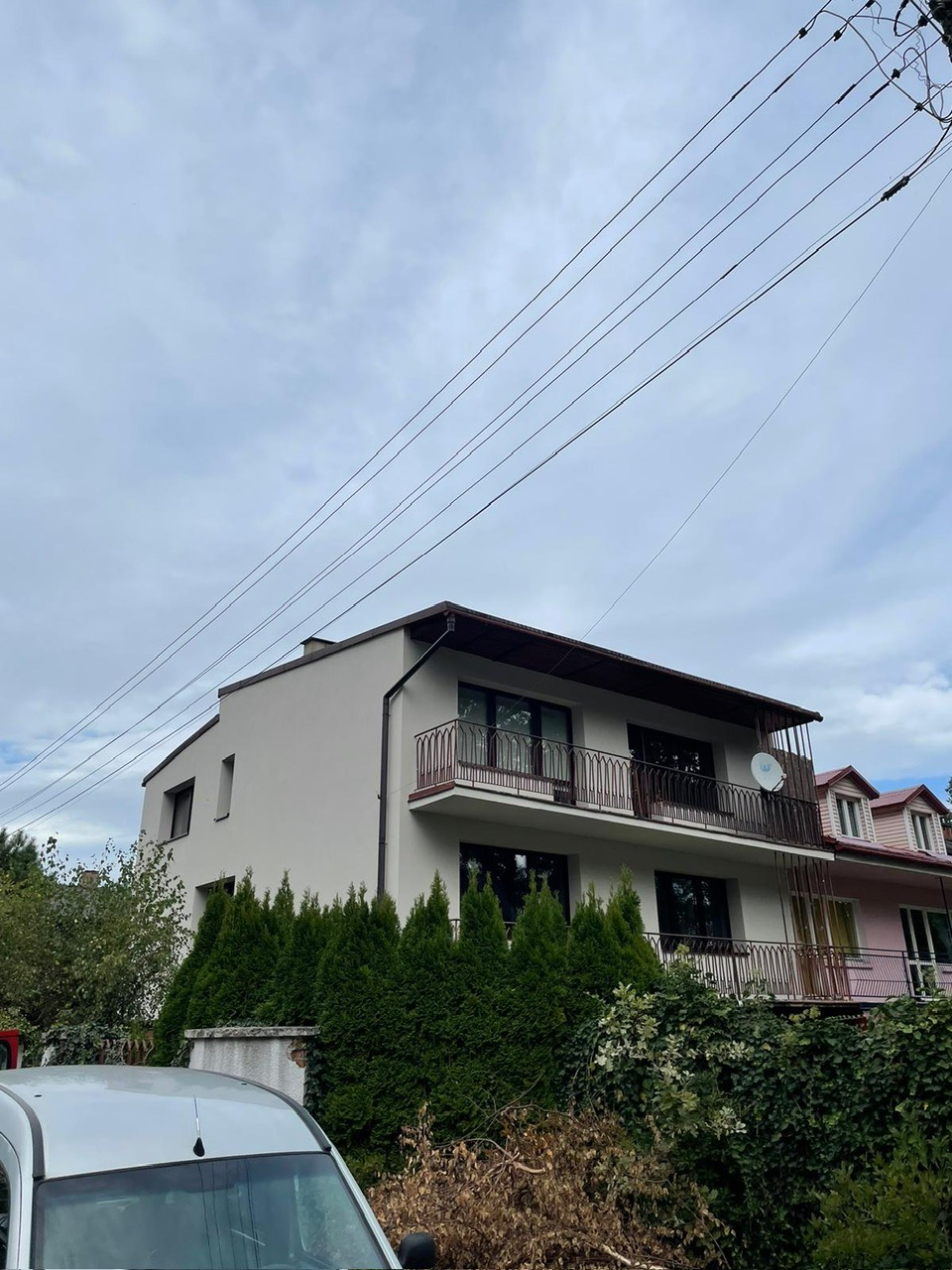 Dwupiętrowy dom z balkonami i jasną elewacją, widok z dołu, częściowo zasłonięty przez żywopłot i zaparkowany samochód dostawczy.