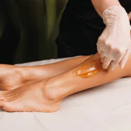 Nakładanie ciepłego wosku na nogę klientki przez kosmetyczkę w białych rękawiczkach, widoczne stopy i zielone rośliny w tle.