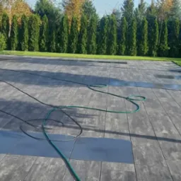 Ułożona, szara kostka brukowa na posesji z zielonym trawnikiem i wysokimi tujami w tle, widoczne ślady wody i zwinięty wąż ogrodowy.