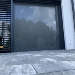 Szara, pionowa przesłona okienna na tle białej ściany budynku, widoczna z poziomu szarego brukowanego tarasu. Po lewej stronie widoczne żaluzje.