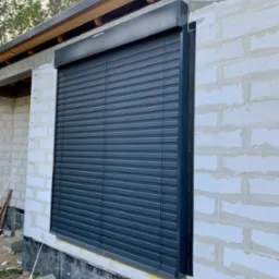 Zamknięta, grafitowa osłona okienna na surowej elewacji z białego gazobetonu, widoczne ślady kleju i montażu, w tle fragment dachu i drzewa.