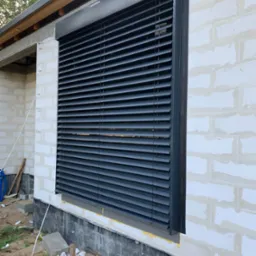 Zamknięte, grafitowe lamele na oknie w surowym budynku z białego bloczka, widoczne elementy konstrukcji dachu, teren wokół okna w trakcie prac wykończeniowych.