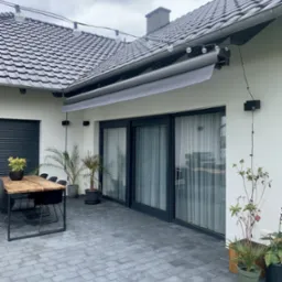 Zewnętrzna szara markiza zwinięta nad tarasem z szarą kostką, drewnianym stołem i czarnymi krzesłami, oświetlonym girlandą żarówek, w tle dom z czarną stolarką okienną i roletami.