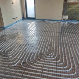 Białe rury ogrzewania podłogowego zamocowane do czarnej siatki na podłodze w trakcie instalacji, widoczne surowe ściany i okno.