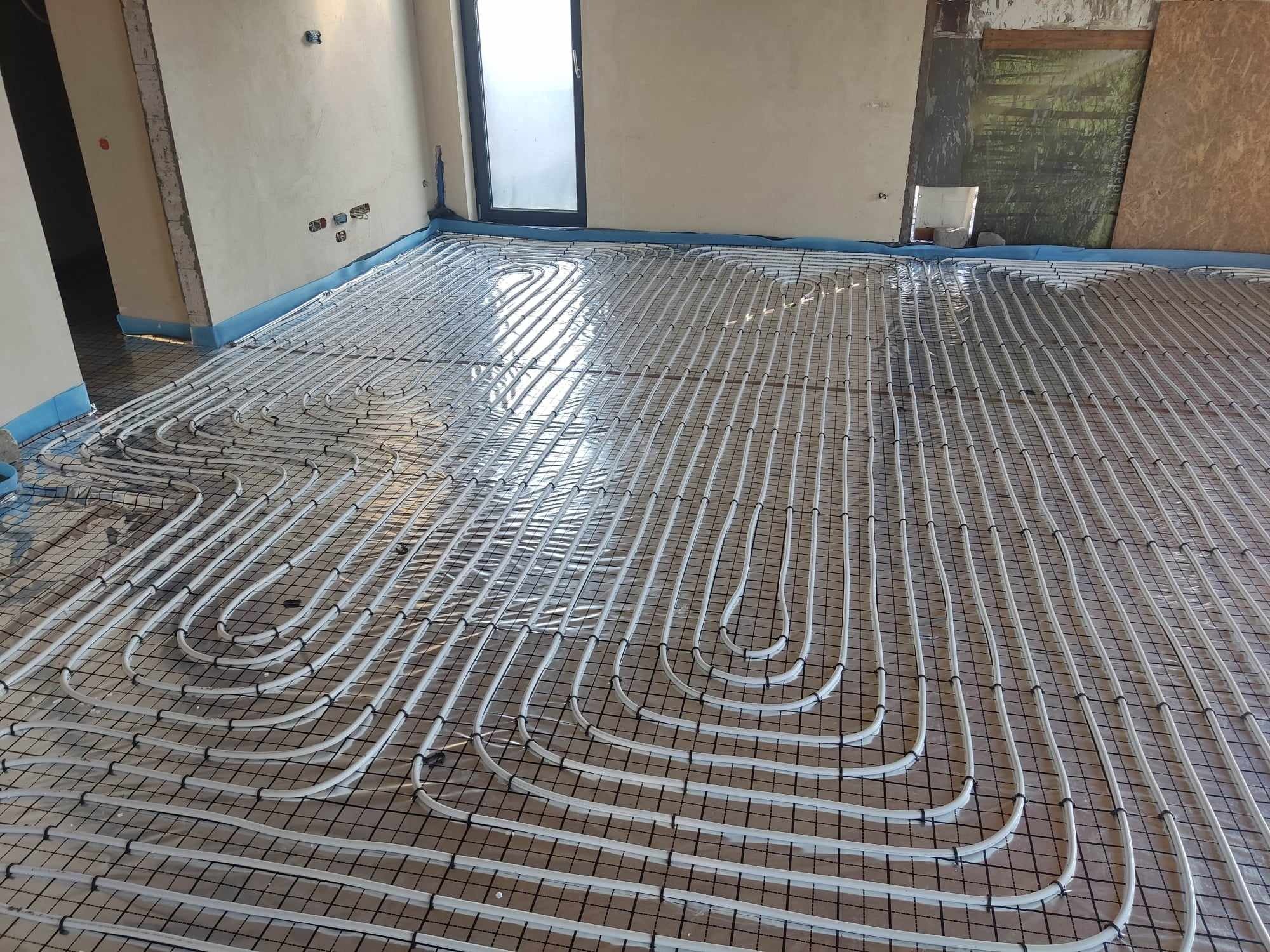 Białe rury ogrzewania podłogowego zamocowane do czarnej siatki na podłodze w trakcie instalacji, widoczne surowe ściany i okno.