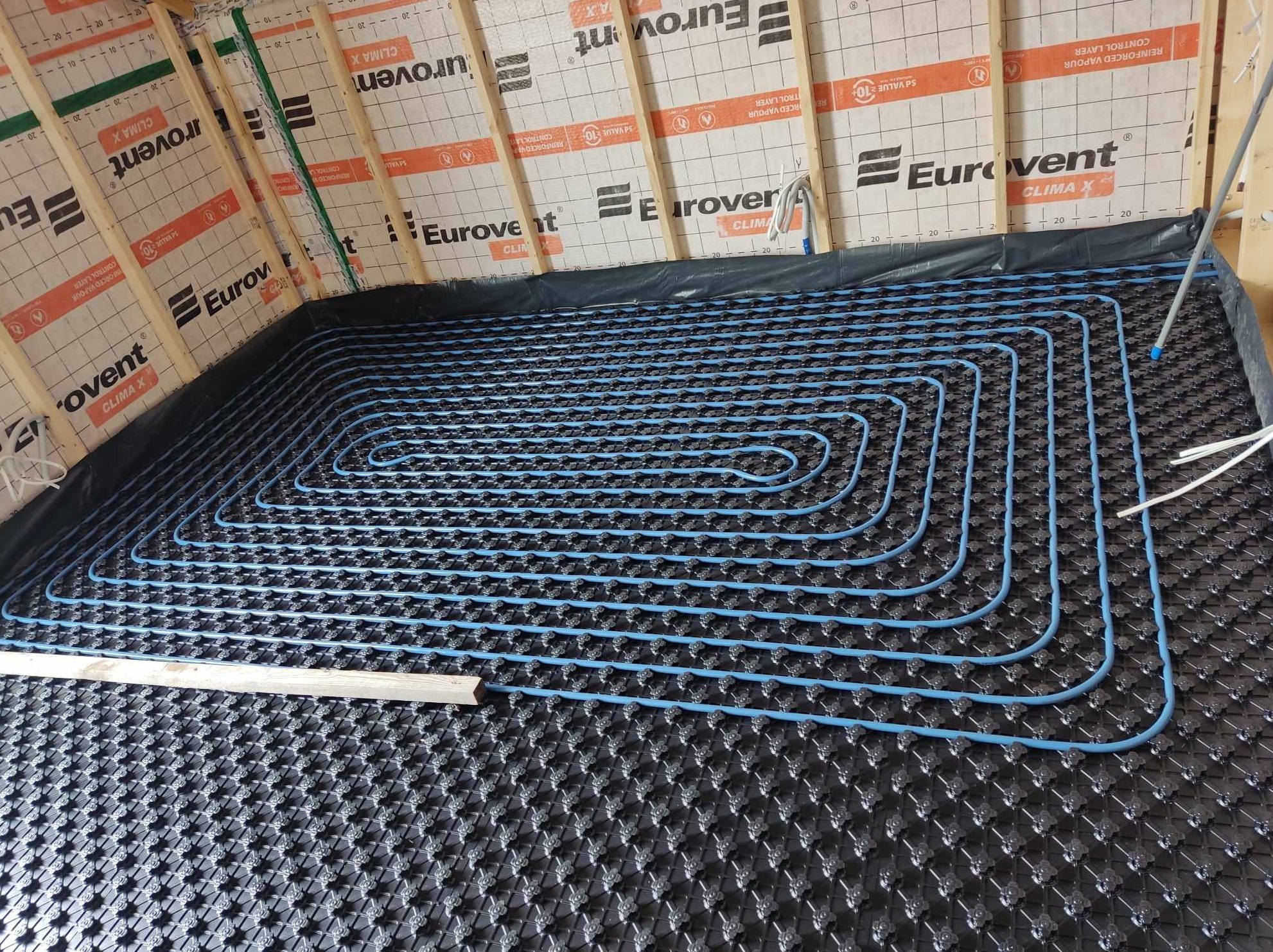 Montaż ogrzewania podłogowego: niebieskie rurki ułożone spiralnie na czarnej macie izolacyjnej z wypustkami, widoczne drewniane elementy konstrukcji dachu i folia paroizolacyjna z nadrukiem Eurovent.