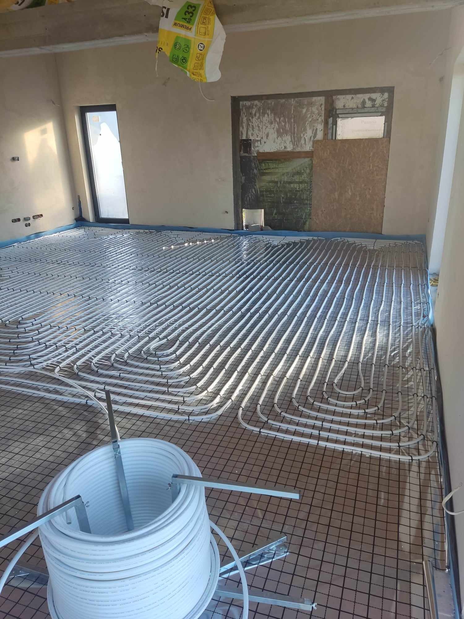 Wnętrze w trakcie instalacji ogrzewania podłogowego: białe rury ułożone na siatce, rolka rur w tle, widać niedokończone ściany i okno.