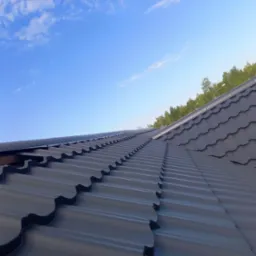 Nowy, grafitowy dach z blachodachówki modułowej, widoczny pod kątem, na tle błękitnego nieba i zieleni drzew.