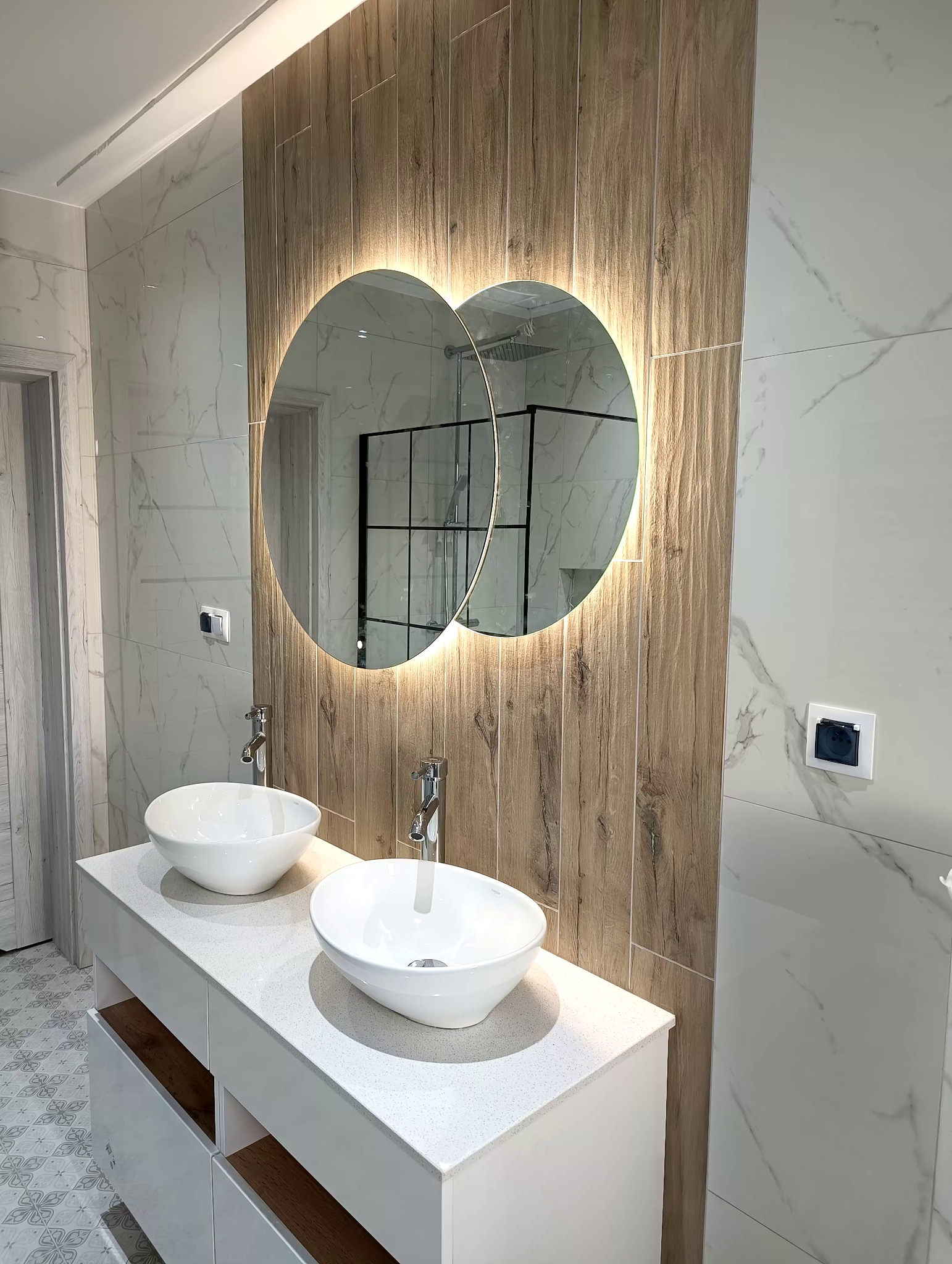 Nowoczesna łazienka z podwójną umywalką nablatową, okrągłym lustrem z podświetleniem LED, drewnopodobnymi i marmurowymi płytkami ściennymi oraz kabiną prysznicową w tle.