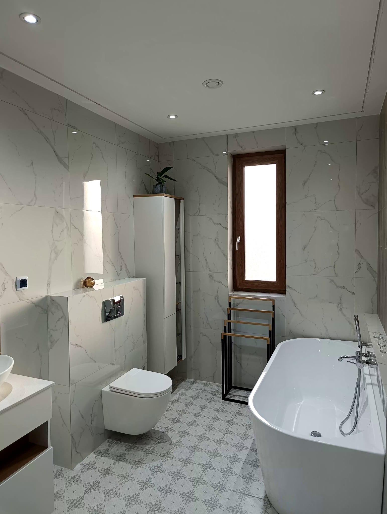 Nowoczesna łazienka z wanną wolnostojącą, wiszącą toaletą, marmurowymi płytkami i wzorzystą podłogą, oświetlona punktowymi lampami sufitowymi i naturalnym światłem z okna.