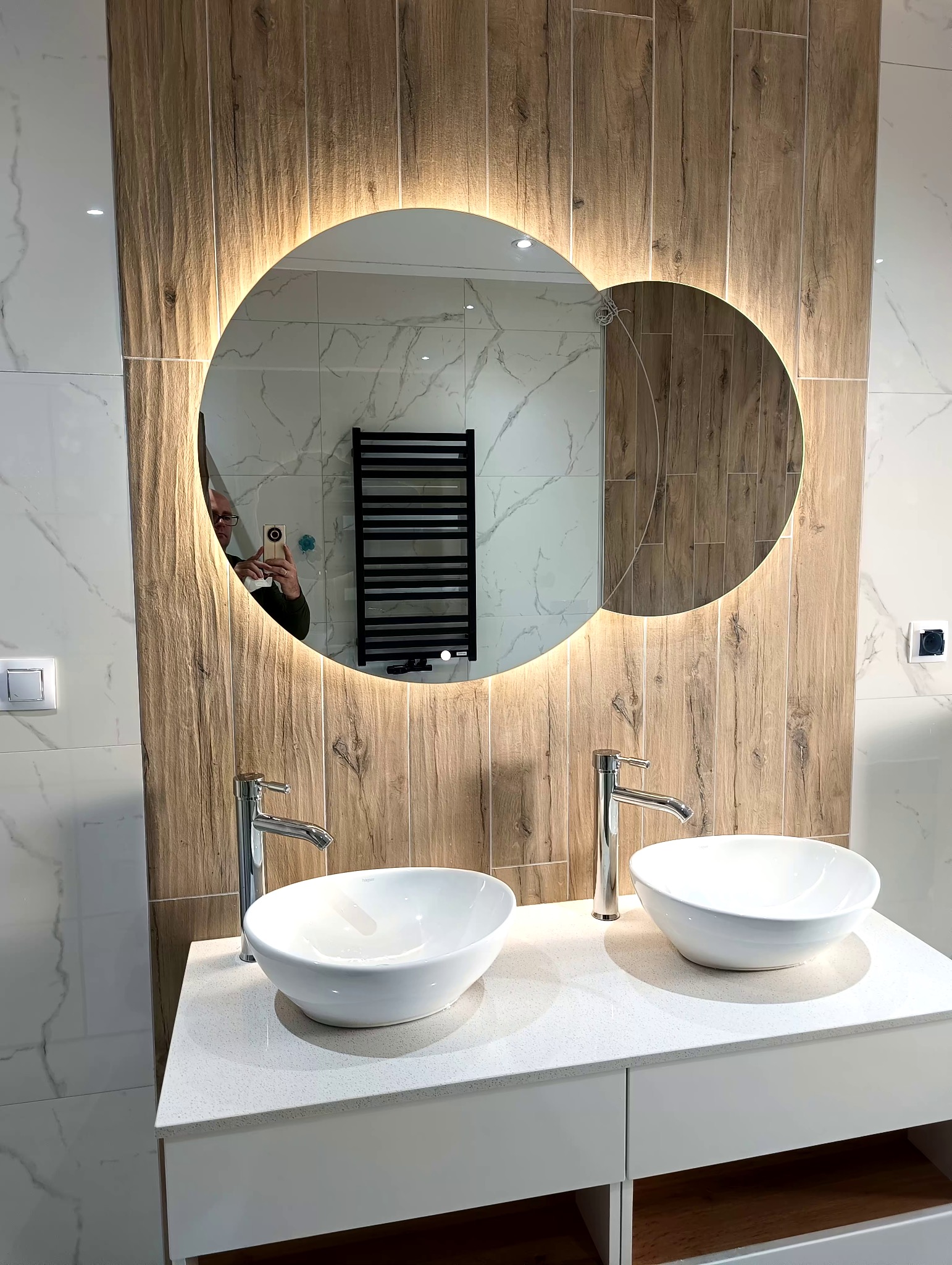 Nowoczesna łazienka z dwoma białymi umywalkami nablatowymi, chromowanymi bateriami i podświetlanym lustrem w kształcie połączonych kół, wykończona płytkami imitującymi drewno i marmur.