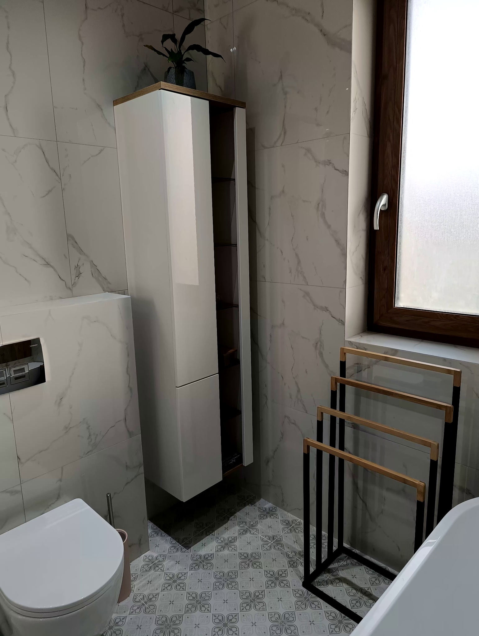 Nowoczesna łazienka z białą szafką wiszącą, marmurowymi płytkami ściennymi i dekoracyjną podłogą w kwiatowy wzór; widoczny fragment wanny i drewniany wieszak na ręczniki.