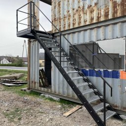 TFS Łukasz Drążek - Zewnętrzne schody metalowe z platformą prowadzące do kontenera z wyciętymi otworami, widoczne elementy konstrukcyjne i otoczenie.