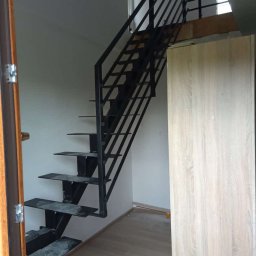 TFS Łukasz Drążek - Wnętrze z metalowymi schodami o czarnej, prostej konstrukcji, z widocznymi stopniami i minimalistyczną balustradą. Jasne ściany i podłoga, drzwi po lewej stronie.