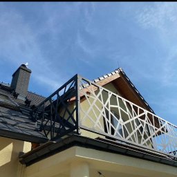 TFS Łukasz Drążek - Czarna, ażurowa balustrada balkonowa z motywem geometrycznym w Głogowie. Widok z dołu na tle błękitnego nieba i dachu domu. Nowoczesny design.