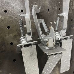 TFS Łukasz Drążek - Dwa srebrne, metalowe pedały z sprężynami na perforowanym stole spawalniczym. Widoczne ślady obróbki i spawania. Detal konstrukcyjny z bliska.