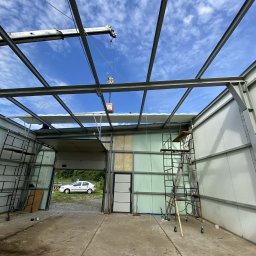 TFS Łukasz Drążek - Wnętrze hali w budowie z widoczną stalową konstrukcją dachu i ścianami z płyt warstwowych. Na pierwszym planie rusztowania i podnośnik. W tle błękitne niebo.