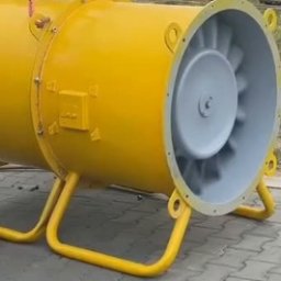 TFS Łukasz Drążek - Żółty, przemysłowy wentylator osiowy na metalowej podstawie, z widocznymi łopatkami wirnika, zawieszony na haku dźwigu, gotowy do montażu w hali.