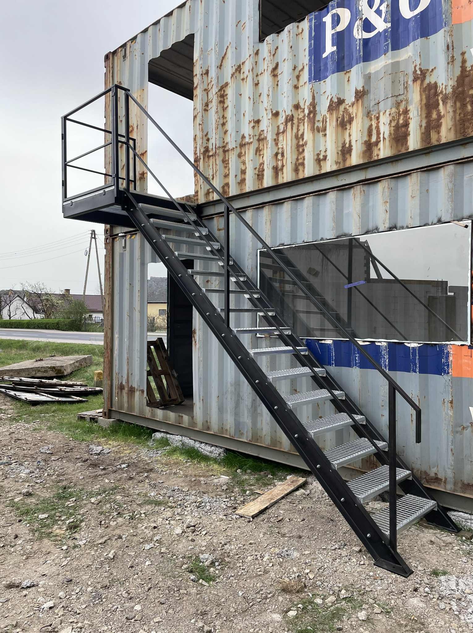 Zewnętrzne schody metalowe z platformą prowadzące do kontenera z wyciętymi otworami, widoczne elementy konstrukcyjne i otoczenie.