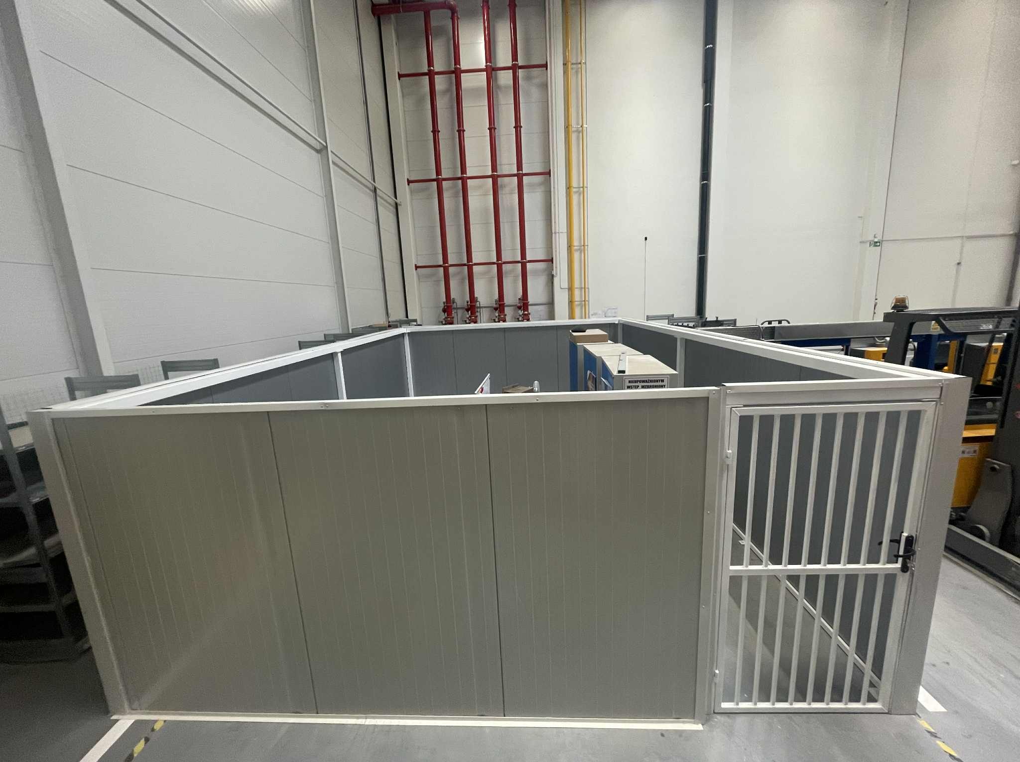 Wykonana z metalu, modułowa konstrukcja w hali produkcyjnej, z kratową furtką. Widoczne czerwone rury instalacji pod sufitem. Szara podłoga i ściany hali.