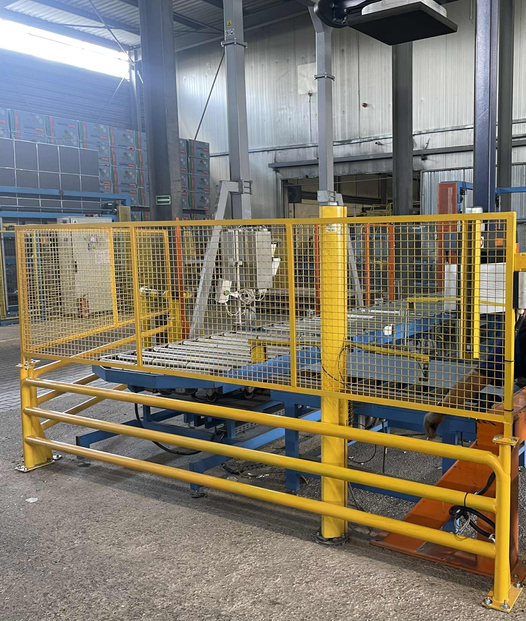 Żółta, metalowa osłona zabezpieczająca maszynę w hali produkcyjnej. Konstrukcja z siatki i rur, niebieska podstawa. Widoczne elementy maszyny i instalacje.