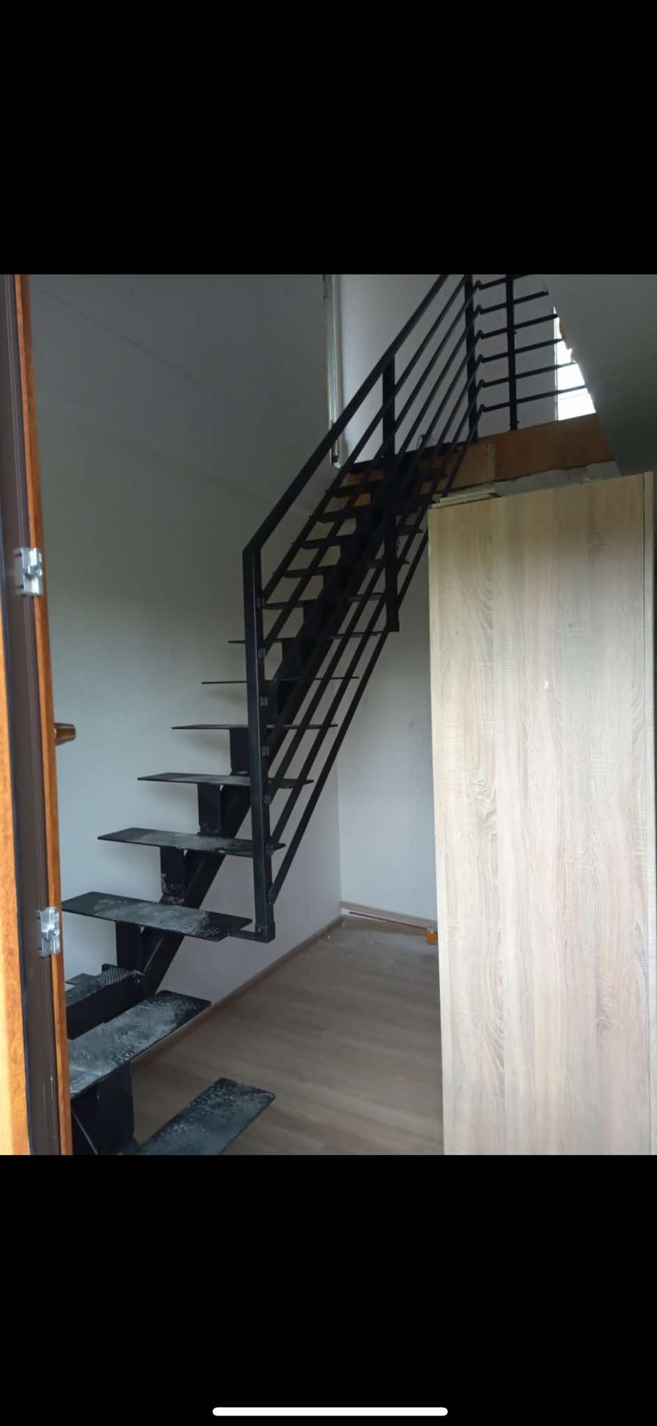 Wnętrze z metalowymi schodami o czarnej, prostej konstrukcji, z widocznymi stopniami i minimalistyczną balustradą. Jasne ściany i podłoga, drzwi po lewej stronie.