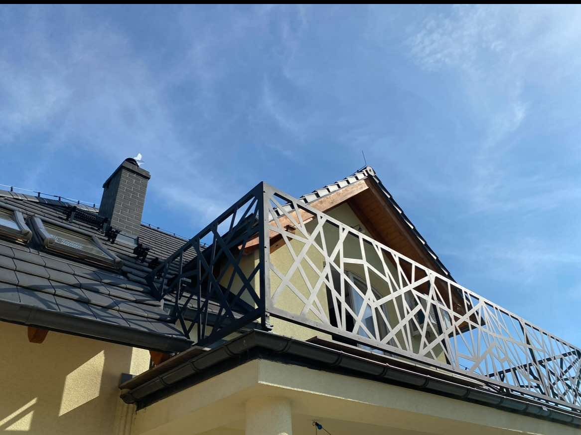 Czarna, ażurowa balustrada balkonowa z motywem geometrycznym w Głogowie. Widok z dołu na tle błękitnego nieba i dachu domu. Nowoczesny design.