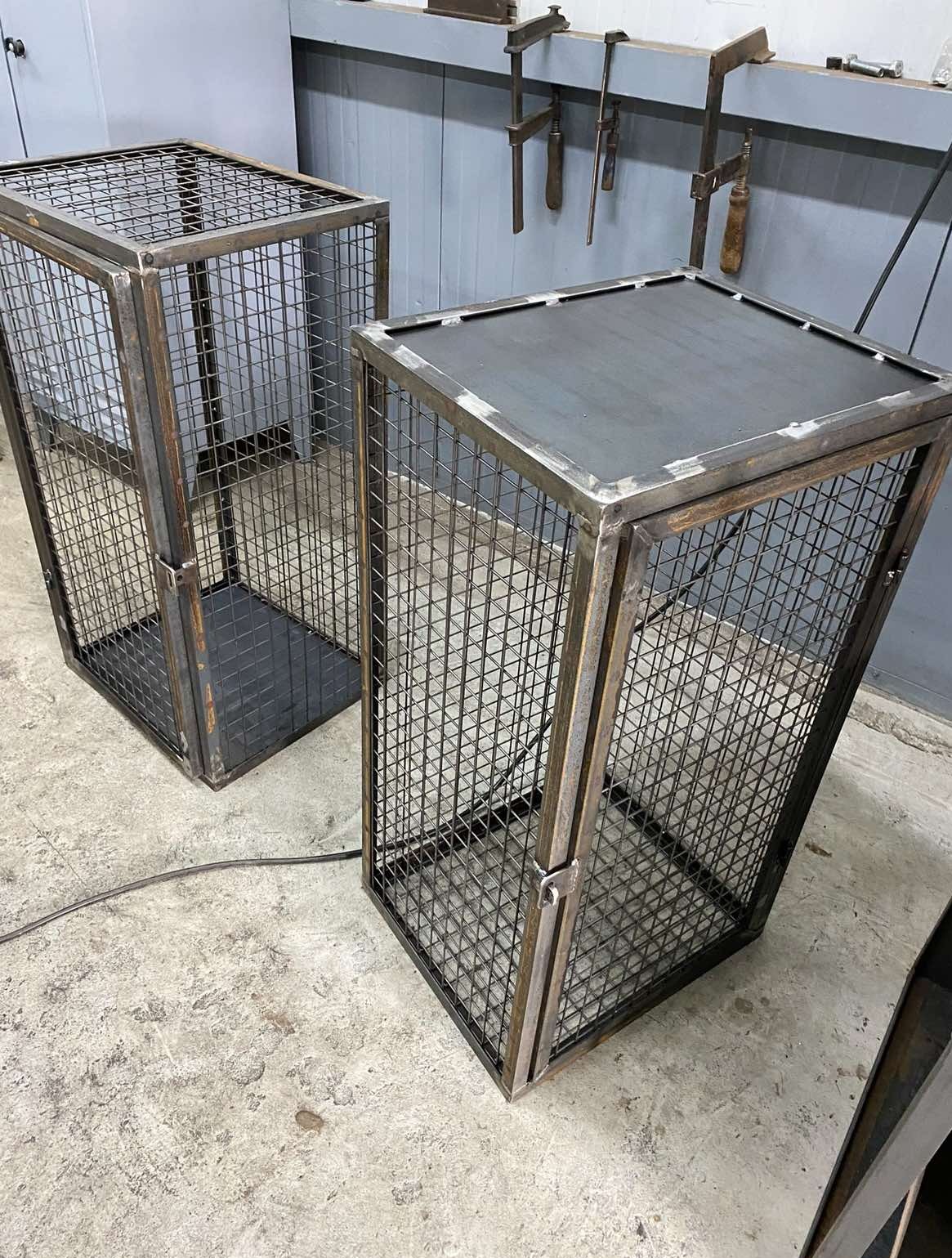 Dwie metalowe klatki o kwadratowej podstawie, wykonane z siatki i stalowych profili, z widocznymi spawami, stojące na betonowej podłodze w warsztacie.