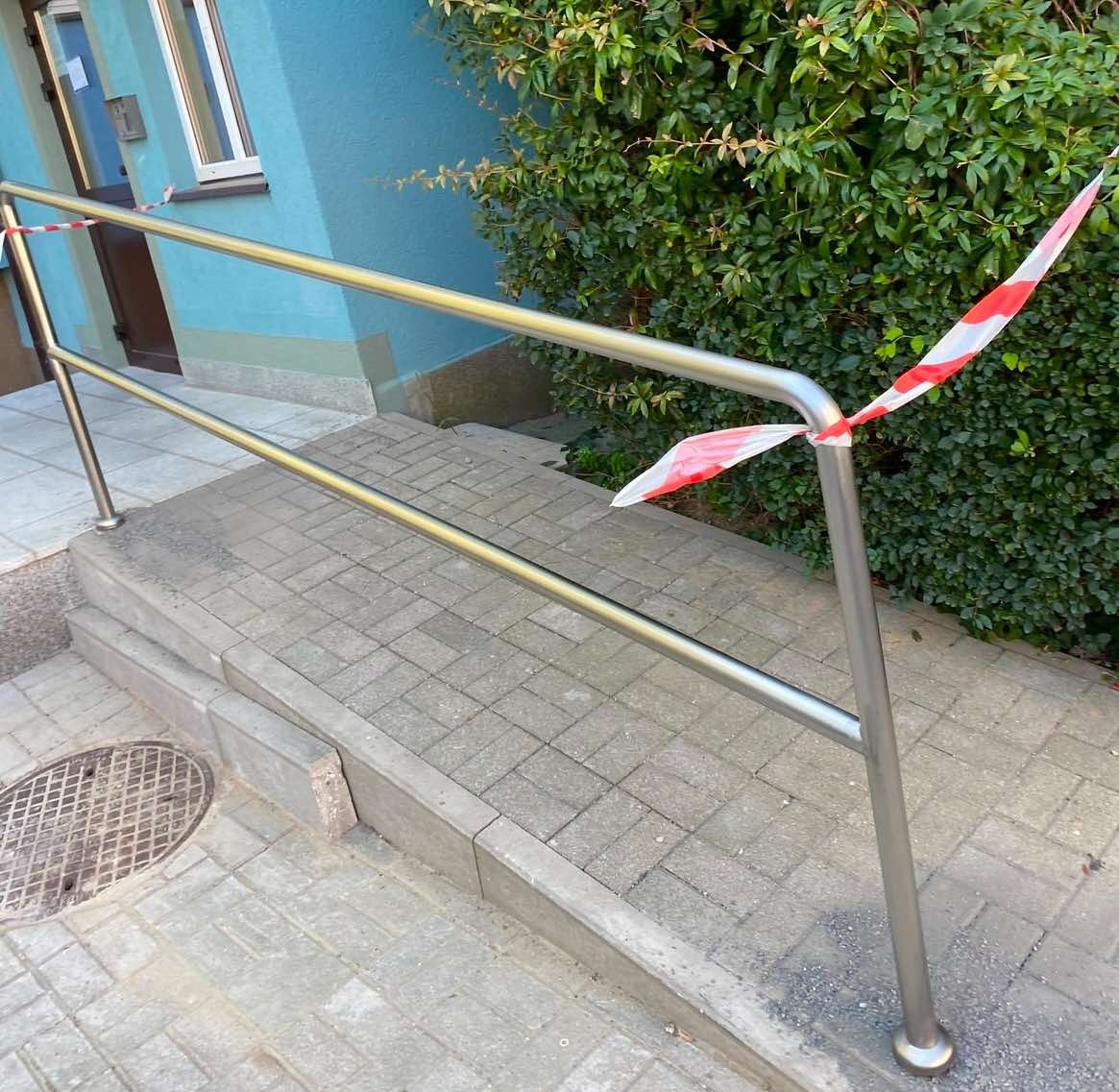 Metalowa poręcz przy schodach wejściowych do budynku, zabezpieczona taśmą ostrzegawczą. Widoczne płytki chodnikowe i fragment zieleni.