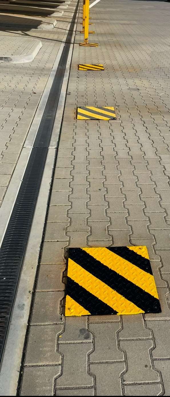 Żółto-czarne, ukośne pasy na kwadratowych płytach chodnikowych, wyznaczające strefę bezpieczeństwa wzdłuż metalowego kanału odwadniającego i słupków.